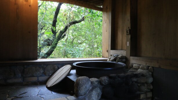 A stone-floored room with a wooden window, a black bathtub, and a stone wall. - Sen-no-ie (Yakushima)