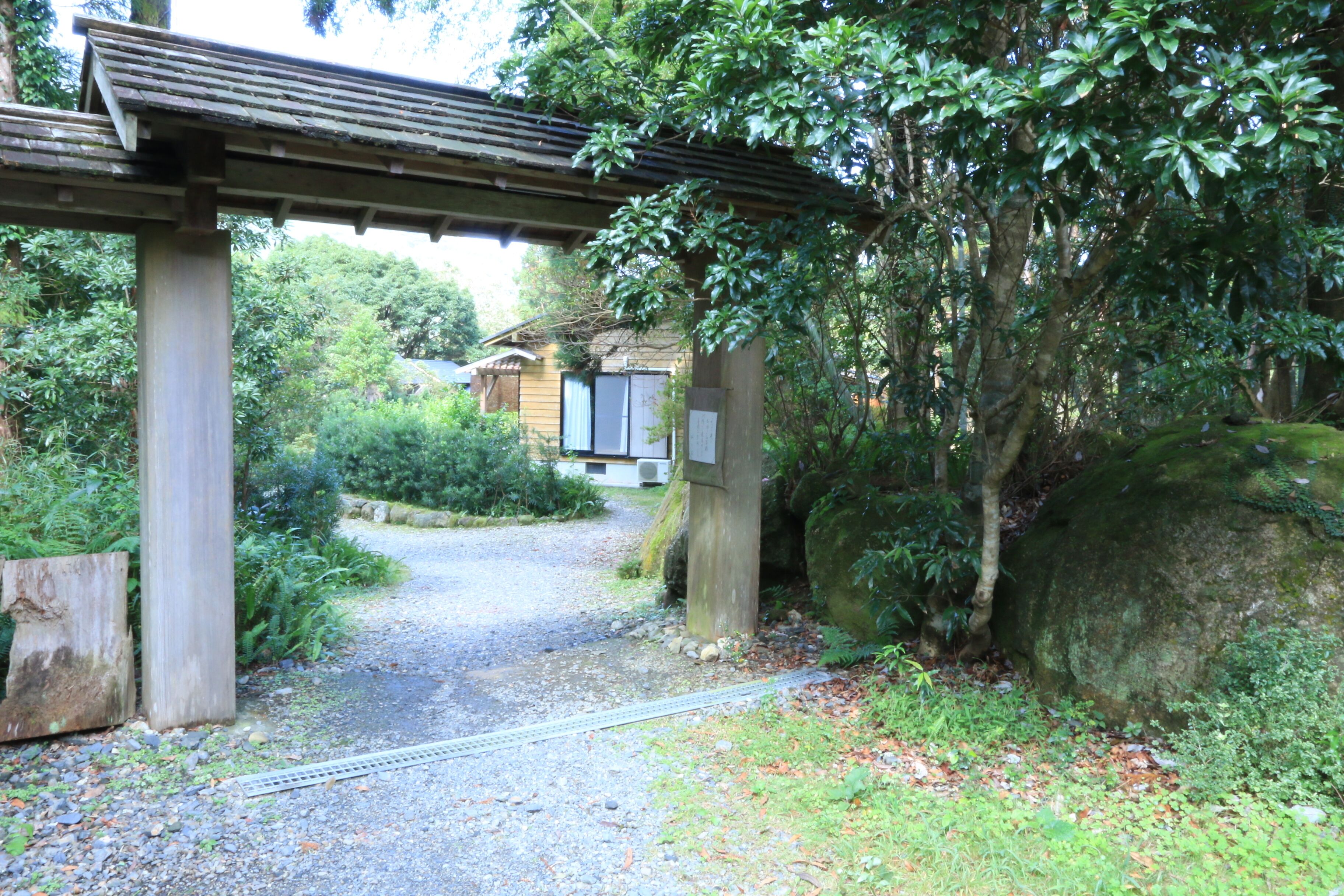 Une passerelle couverte en bois mène à un bâtiment de style architectural japonais traditionnel, entouré d’une végétation luxuriante et bordé d’un chemin de gravier.