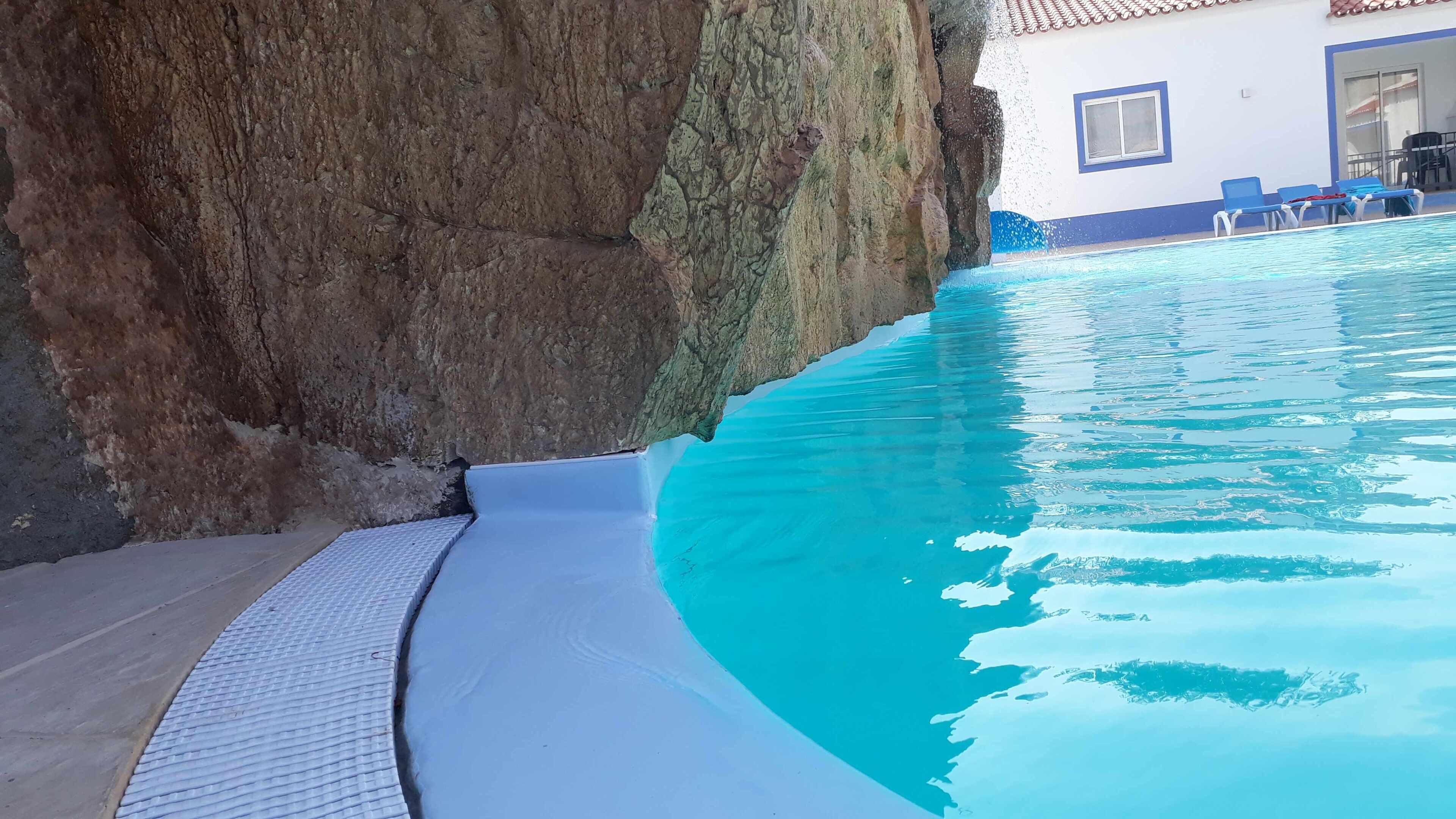 En swimmingpool som ligger intill en klippig brant, med en kakelbeklädd poolterrass och en byggnad i bakgrunden.