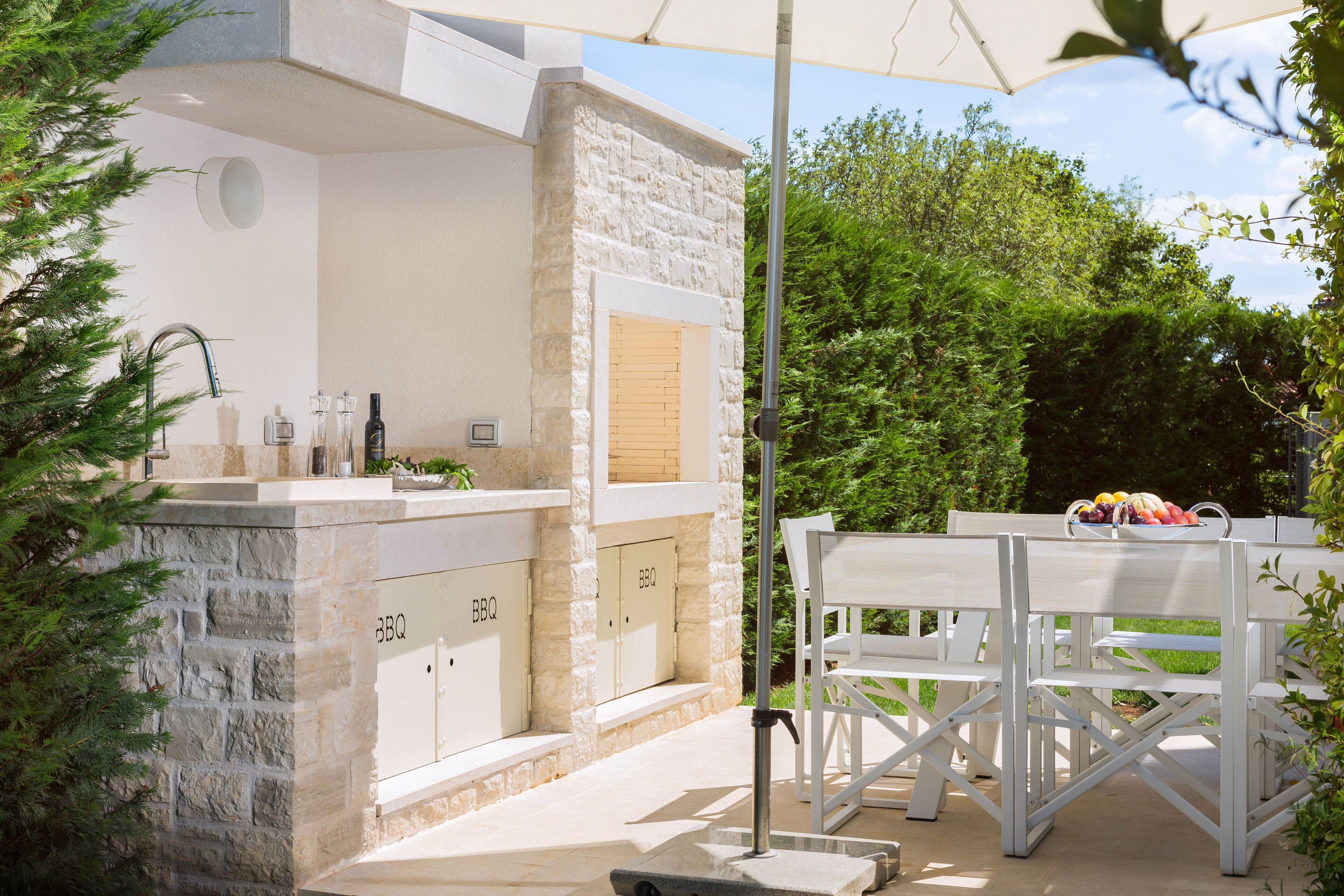 A stone BBQ area with a sink and a wooden door, surrounded by greenery.
