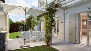 Un patio exterior moderno con una mesa y sillas blancas, un paraguas grande y un árbol pequeño.