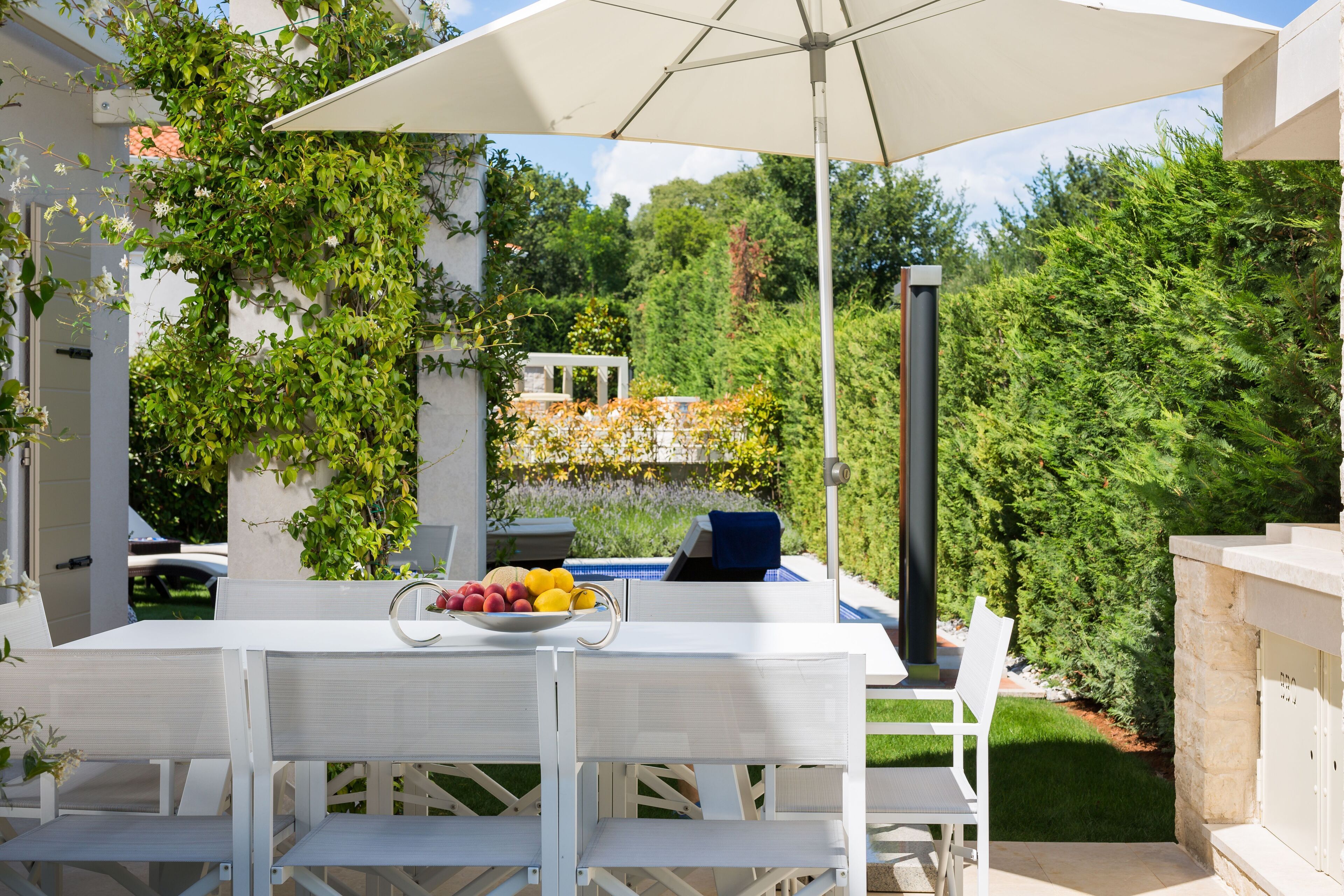 An outdoor patio with a white table and chairs, a bowl of fruit, and a large umbrella.