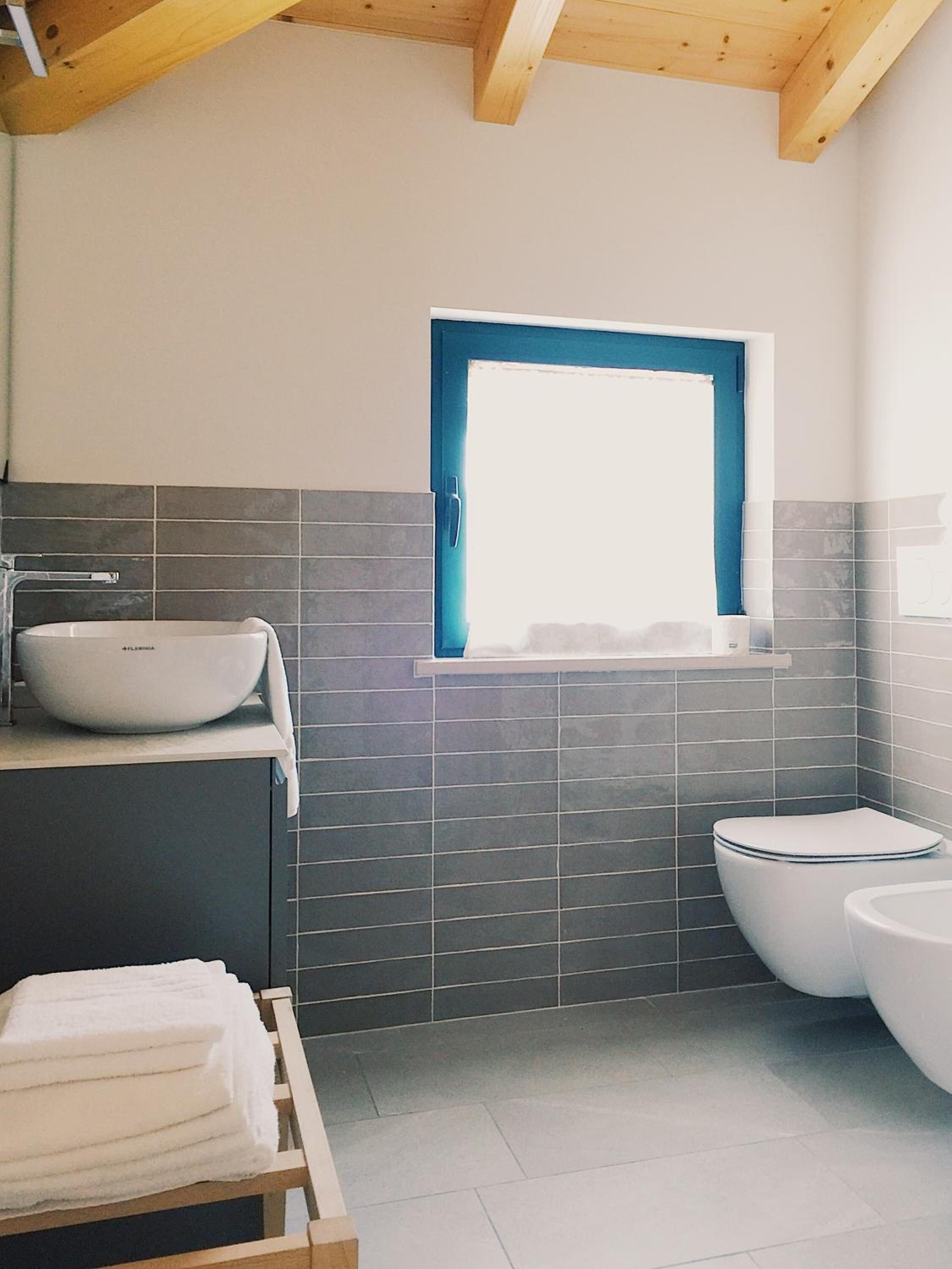 A modern bathroom with a large white sink, a toilet, and a bidet. There is a window with a blue frame and a towel rack with folded towels.