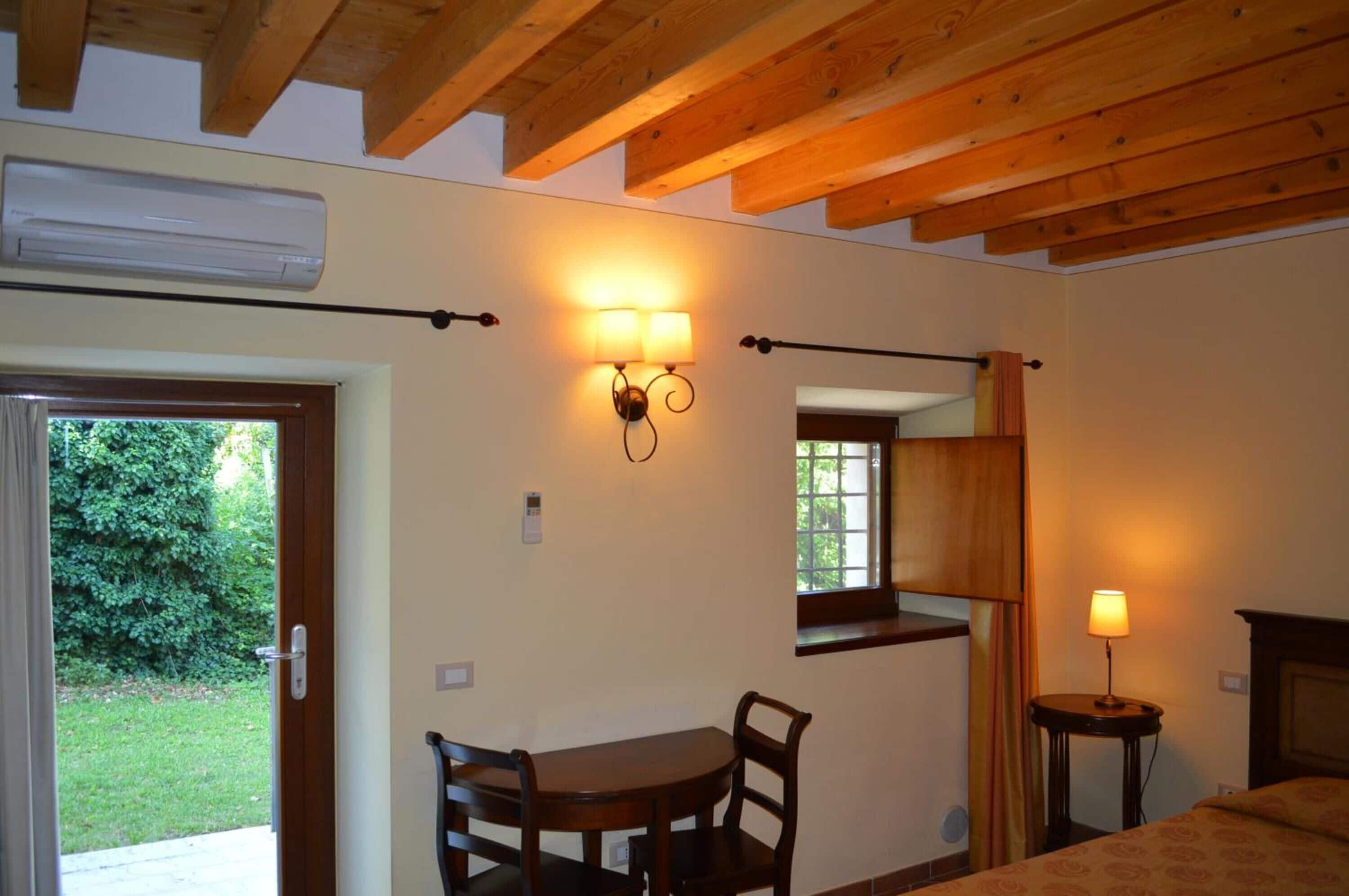 Ein Zimmer mit Holzboden, einem Tisch mit Stühlen, einem Bett, einem Fenster mit Vorhängen und einer Wandleuchte.