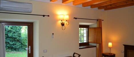 Una habitación con techo de madera, una mesa con sillas, una cama, una ventana con cortinas y una lámpara fijada a la pared.