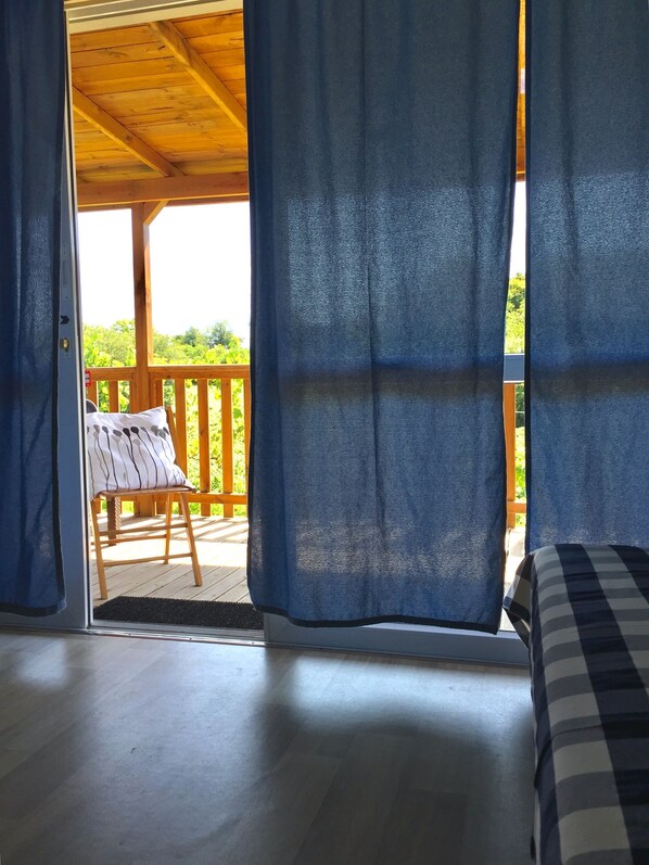 A room with a wooden ceiling, a bed with a checkered bedspread, and a view of an outdoor deck with a chair and railing.