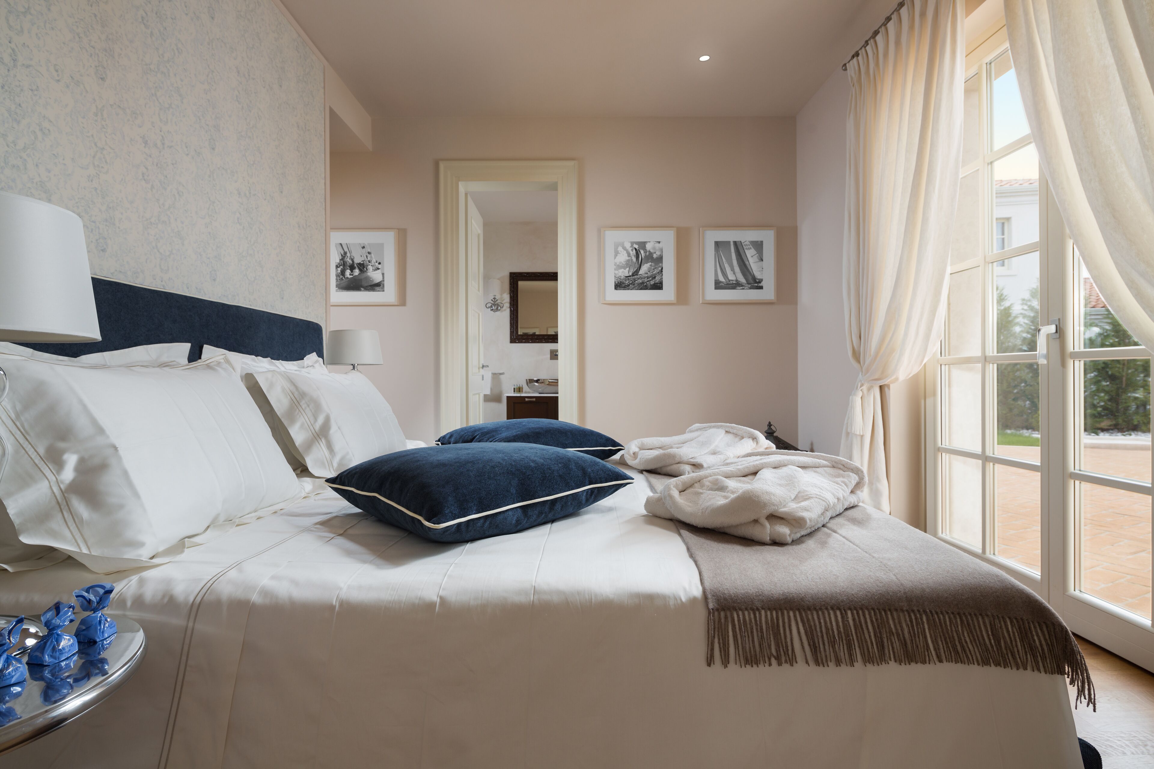 Cama bem arrumada com almofadas brancas e azuis, um cobertor bege e uma mesa de cabeceira com abajur. Há quadros nas paredes e uma janela com cortinas.