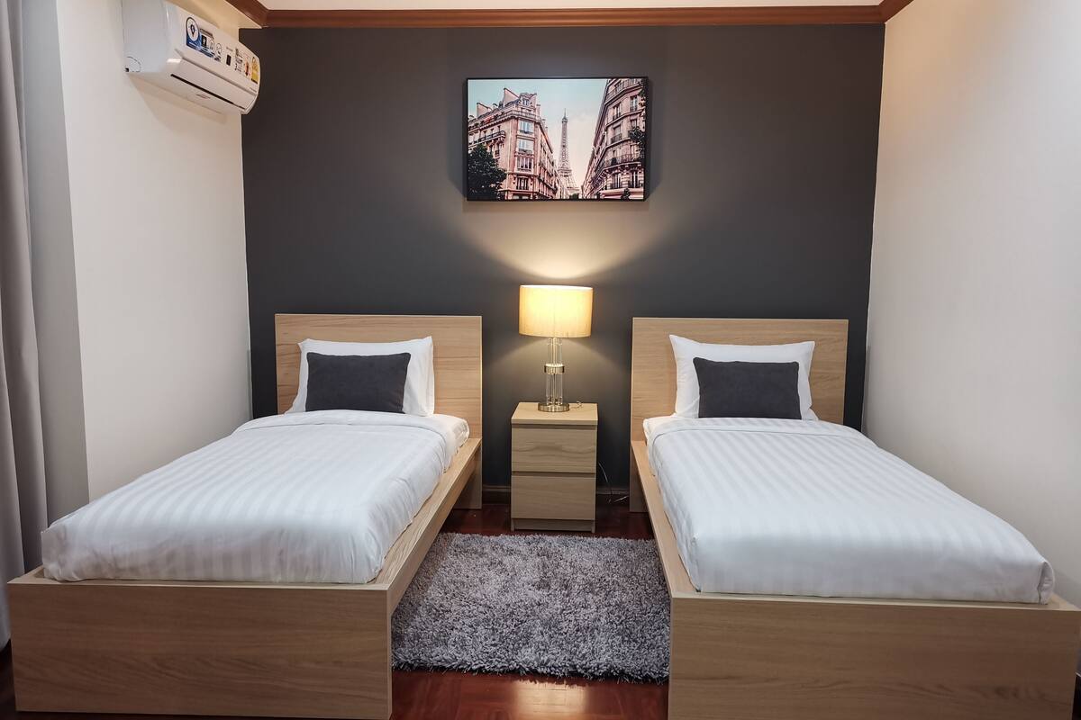 A hotel room with two wooden beds, a grey rug, a nightstand with a lamp, and a picture of Parisian buildings on the wall.
