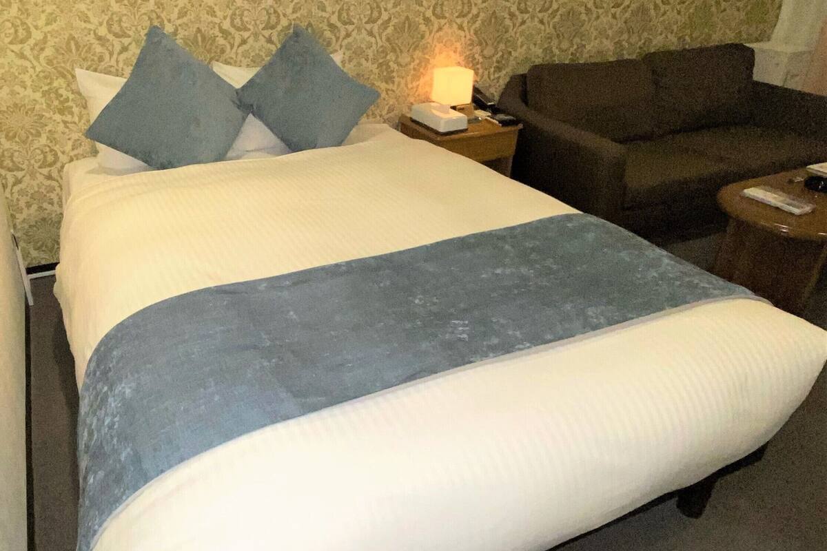 Ein Hotelzimmer mit einem Bett, einem Nachttisch mit Lampe, einer braunen Couch und einem kleinen Tisch mit Fernbedienung.