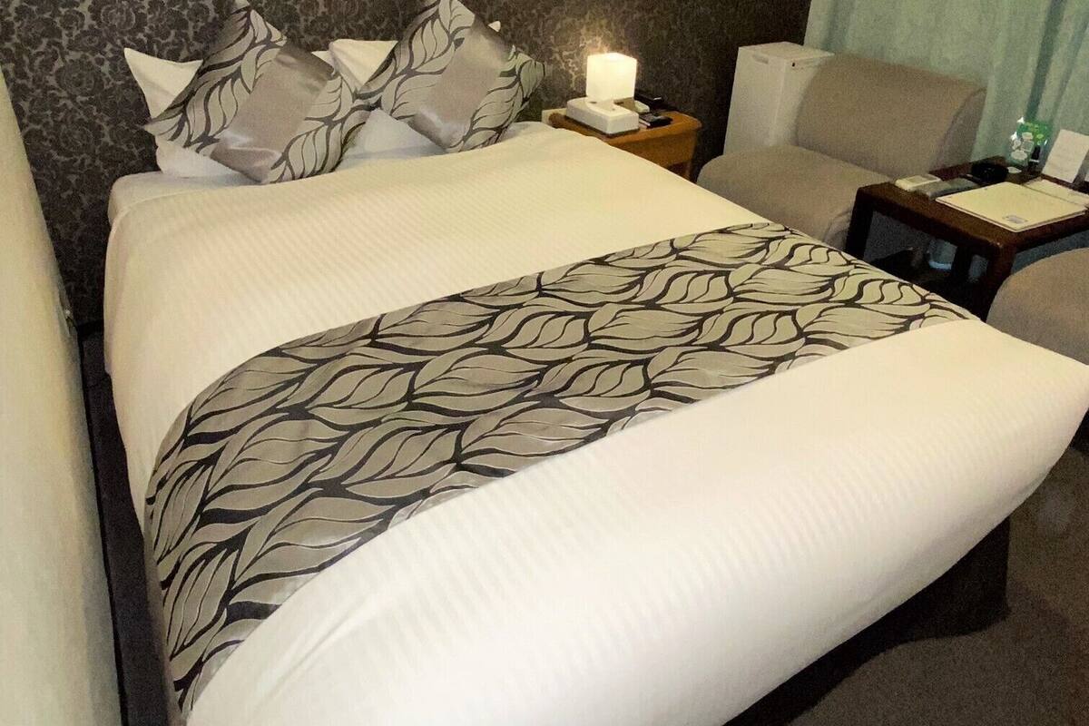 Ein Hotelzimmer mit einem großen Bett, einem Nachttisch mit Lampe, einem Sessel und einem kleinen Tisch mit Telefon und Unterlagen.