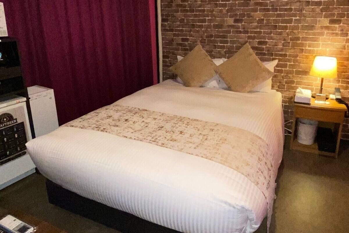 Ein Hotelzimmer mit Backsteinwand, einem Bett mit weißer Bettwäsche, einem roten Vorhang und einem Nachttisch mit Lampe und Taschentuchbox.