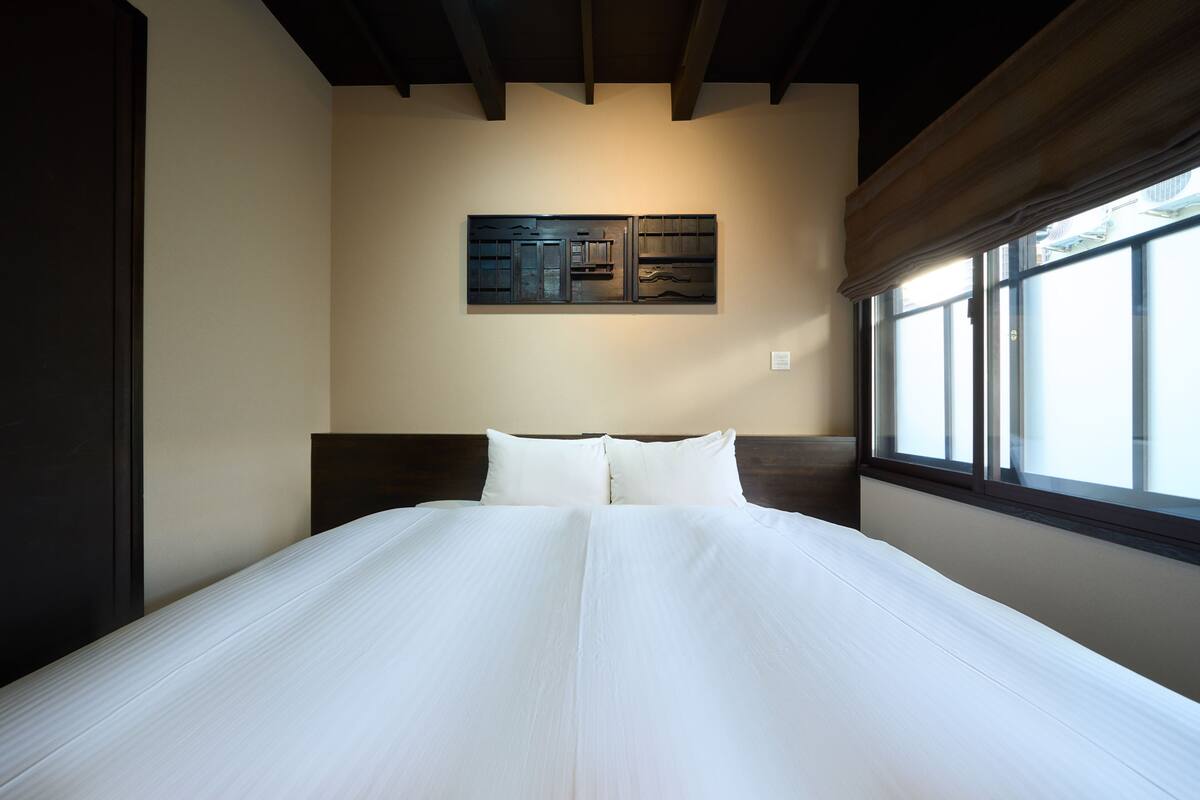 A neatly made bed in a hotel room with a large window, a wall-mounted artwork, and a wooden ceiling.