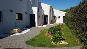 Una casa moderna de dos pisos con entrada de grava, un pequeño jardín con plantas y una roca, y un seto bien recortado.