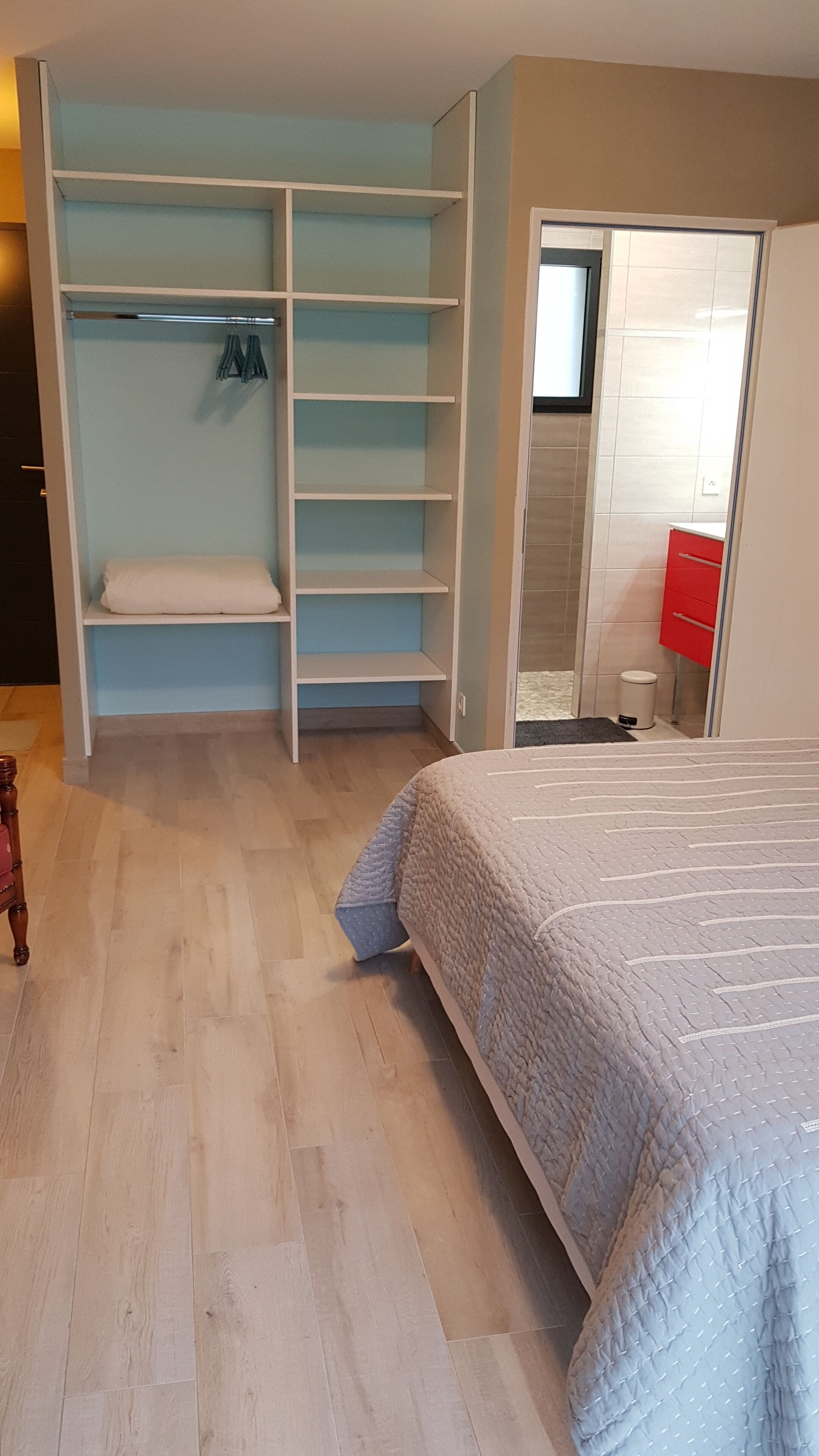 A bedroom with a bed, a wardrobe with multiple shelves, a small stool, and a red cabinet.