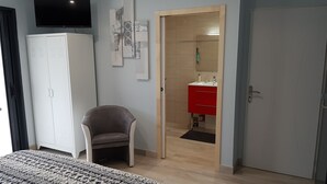 Un dormitorio con cama, armario, silla, televisor y baño visible a través de una puerta abierta.