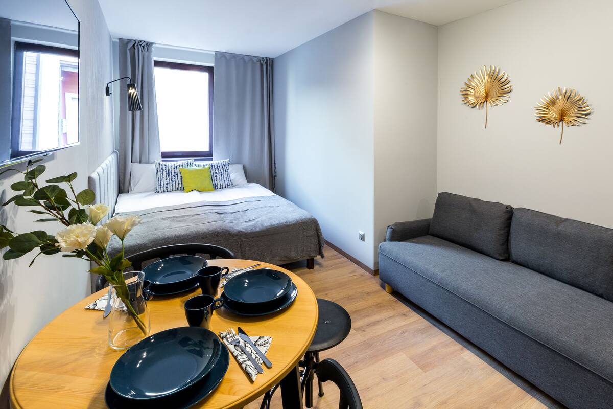A compact living space with a bed, a sofa, a dining table with plates and cutlery, and a vase with flowers.