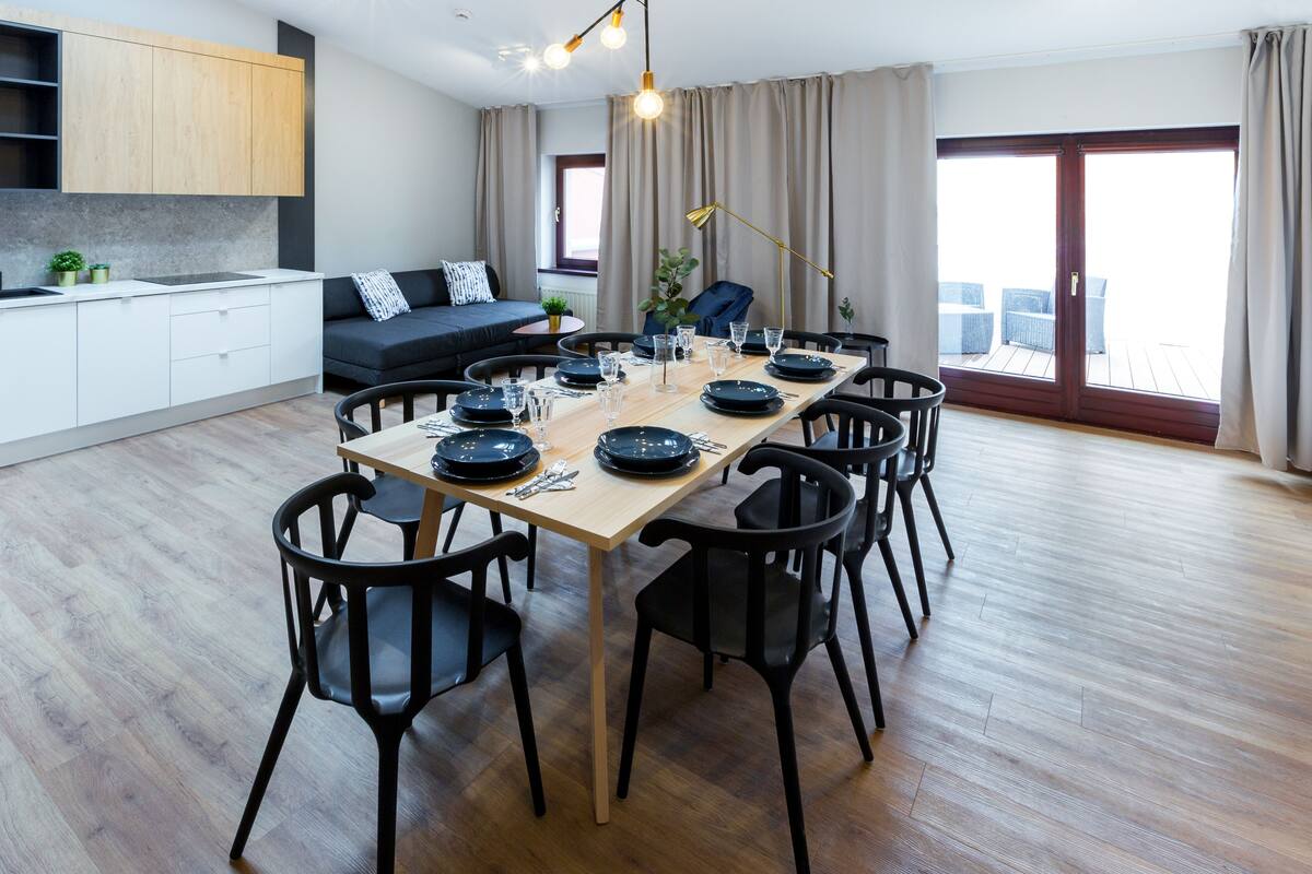 A modern dining area with a wooden table set for six, surrounded by black chairs. A sofa and a small kitchen area are visible in the background.