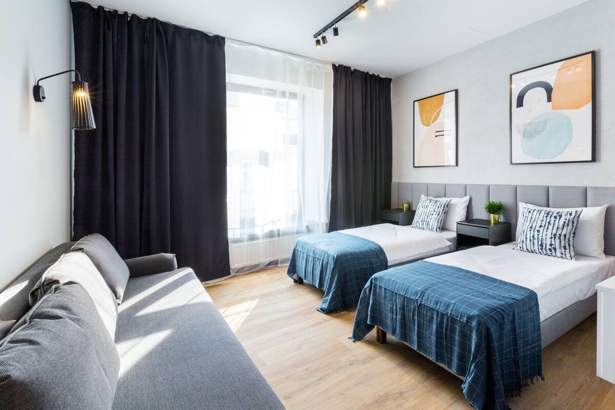 A modern hotel room with two beds, a sofa, a bedside table with a lamp, and artwork on the walls.
