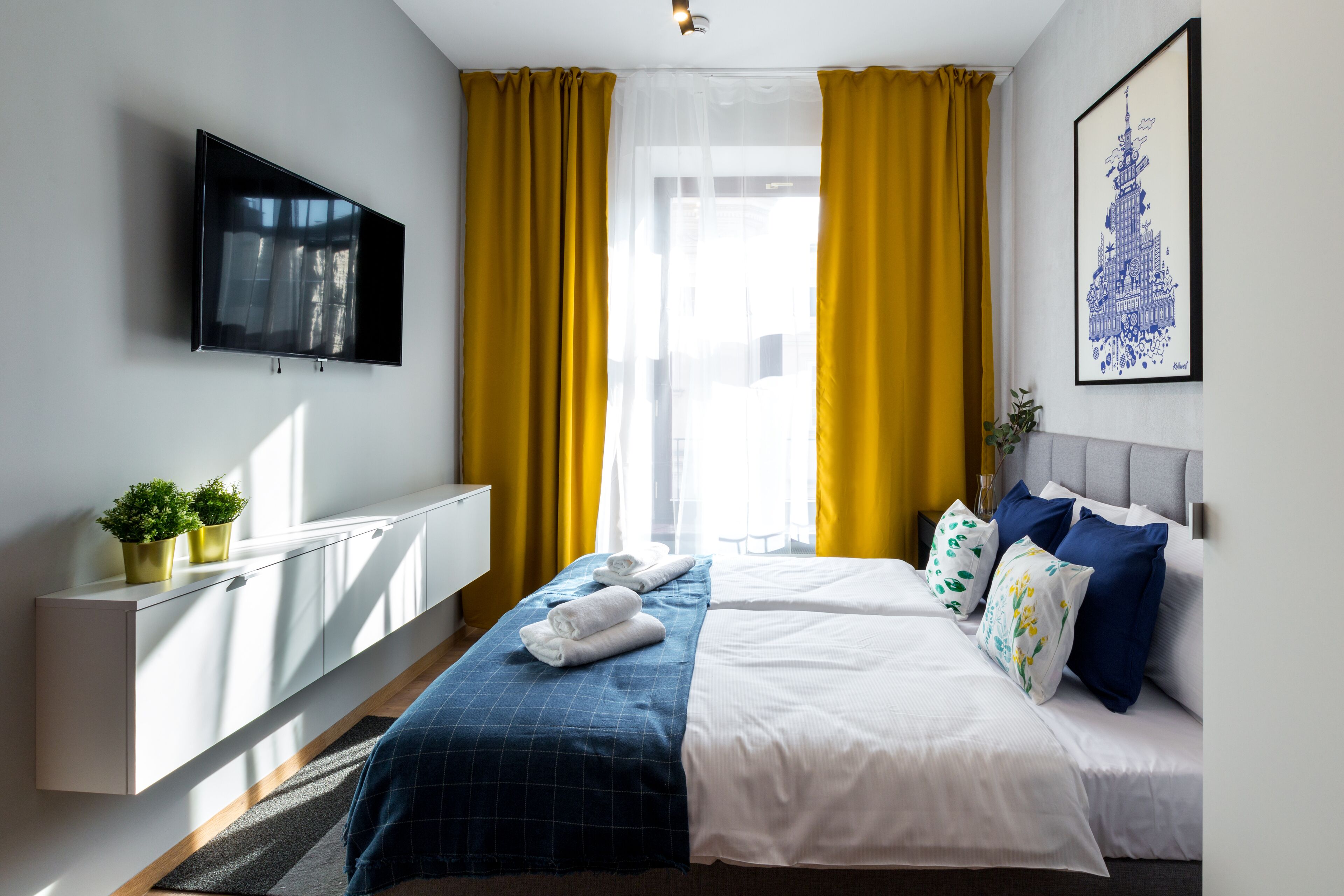 A hotel room with a bed, a TV, a window with yellow curtains, and a wall-mounted artwork.