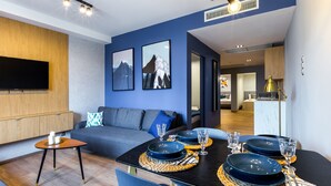 Ein modernes Wohnzimmer mit Essbereich, inklusive Flachbildfernseher, einer Couch mit Kissen und einem Esstisch, der mit Tellern, Gläsern und Besteck gedeckt ist.