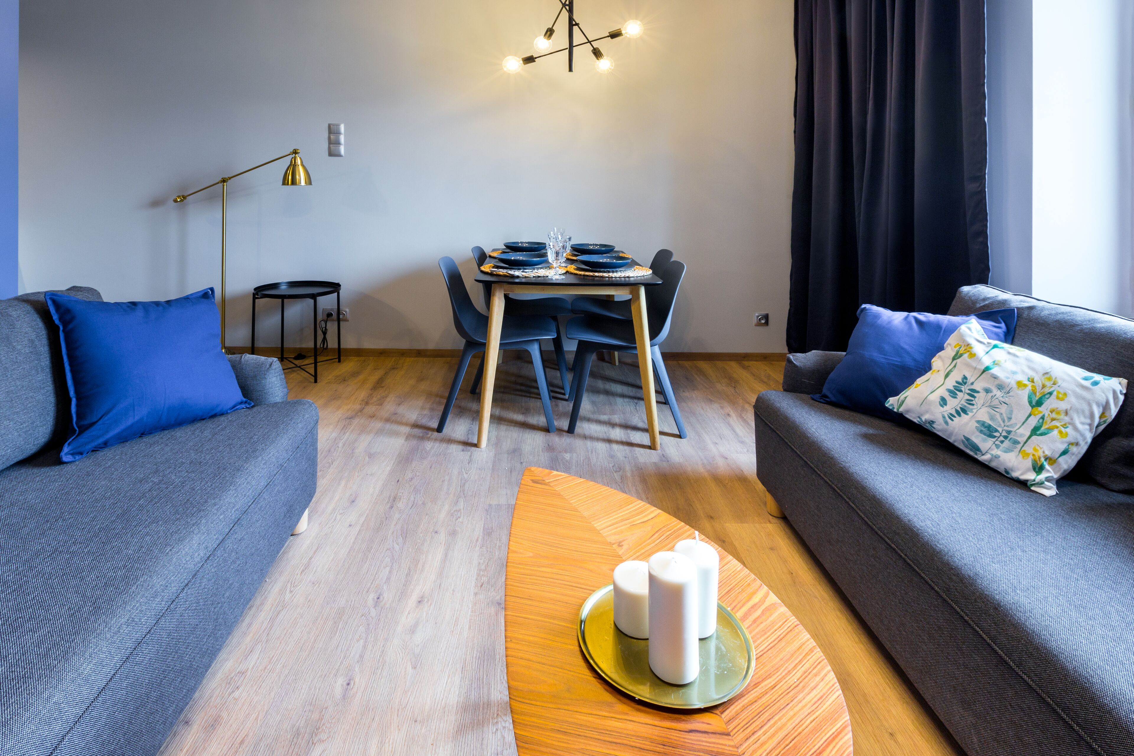 A modern living room with a dining table set for four, two sofas with blue cushions, a floor lamp, and a chandelier.