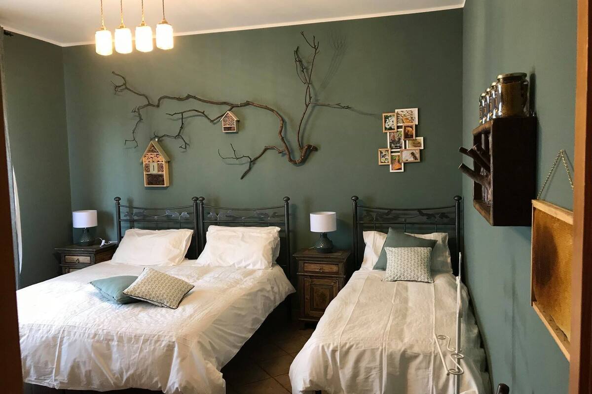Een slaapkamer met twee bedden, elk met een nachtkastje en een lamp. Er zijn decoraties aan de muur, waaronder een tak, een vogelhuisje en een verzameling ingelijste foto’s.