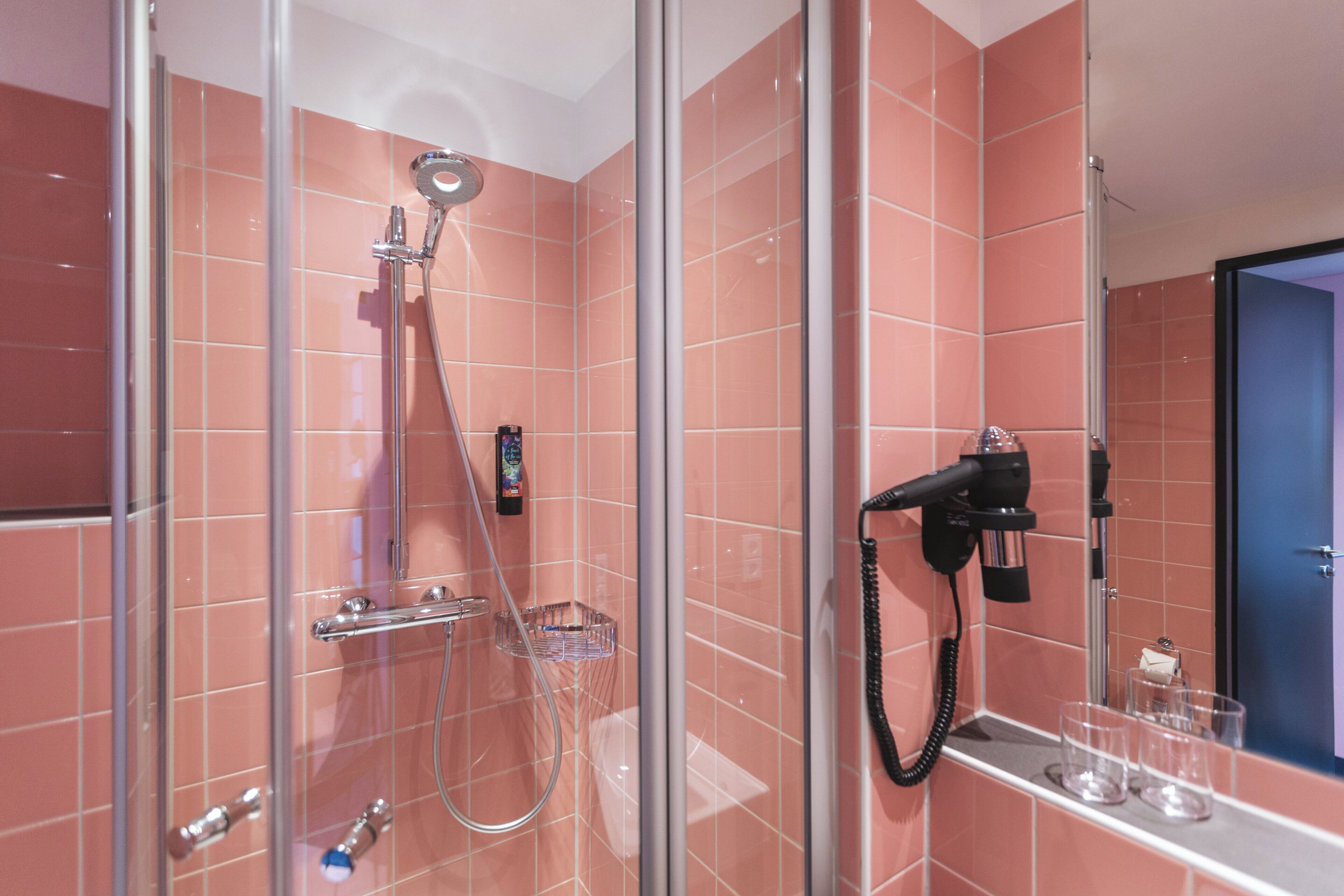 Ein modernes Badezimmer mit rosa Kacheln, einer gläsernen Duschkabine, einem Föhn auf einer Ablage und einem kleinen Regal mit Gläsern.
