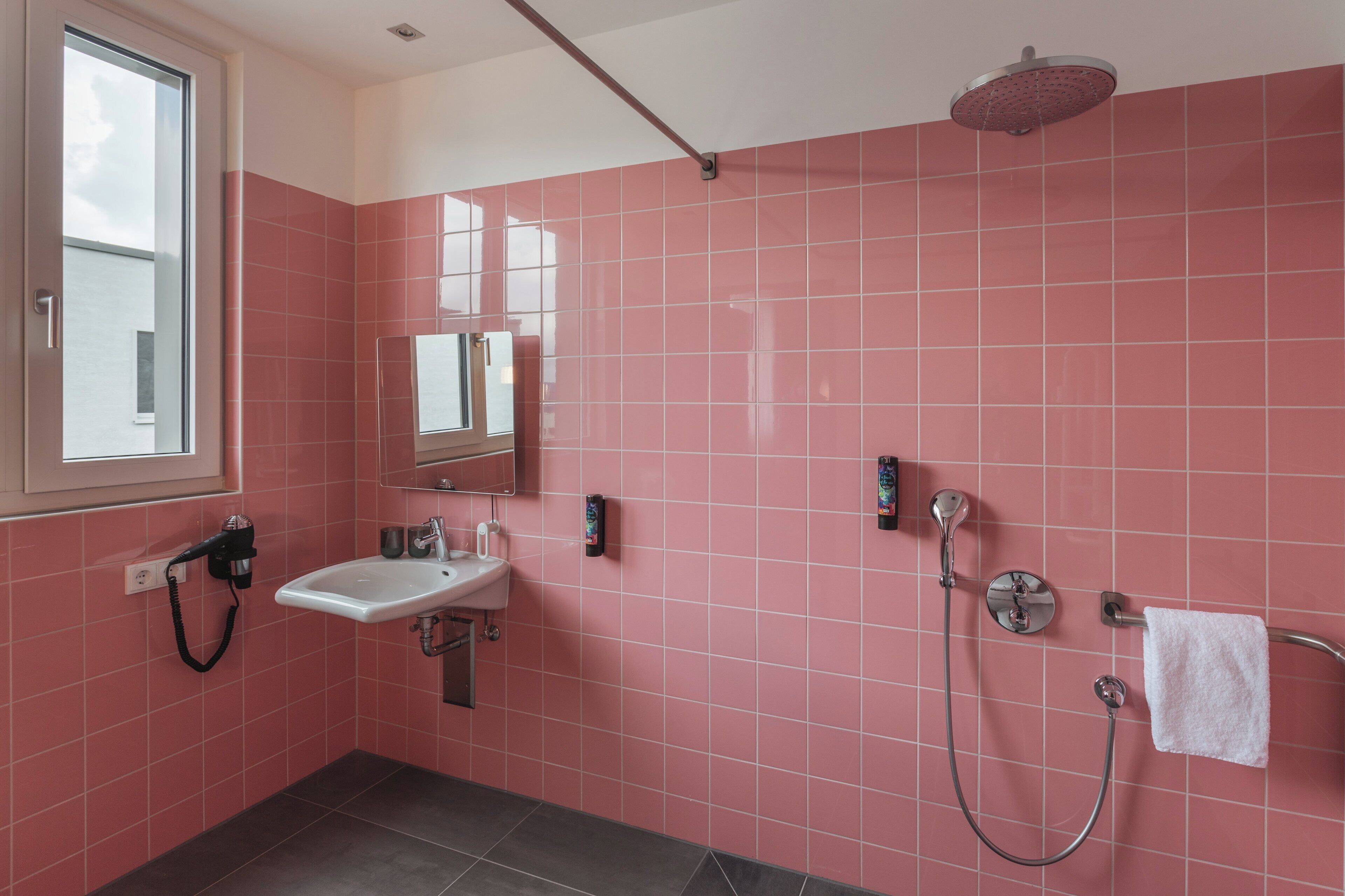 Un baño con azulejos rosa, un lavabo, un espejo, una ducha y un perchero para toallas.