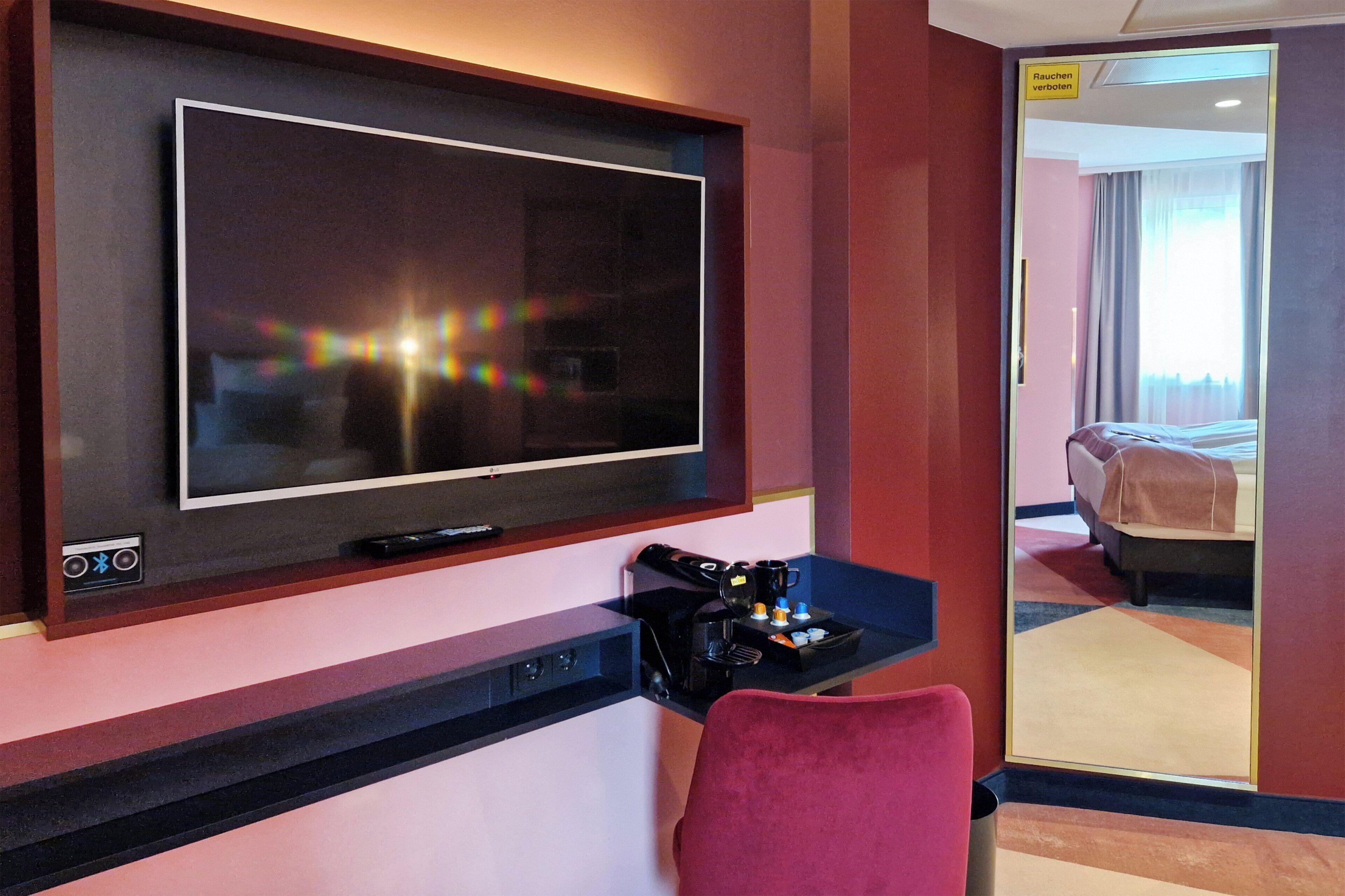 Ein Hotelzimmer mit Flachbildfernseher an der Wand, einer Kaffeemaschine und einem kleinen Tisch mit Fernbedienung.