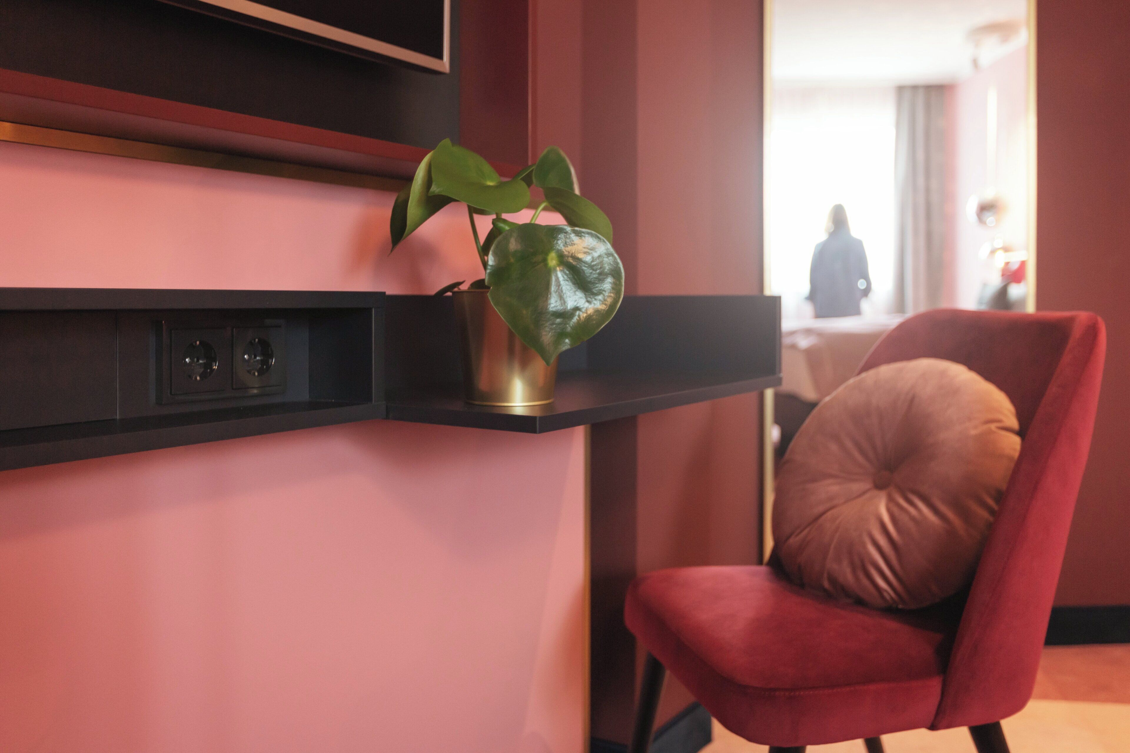 Un lobby de hotel moderno con una silla roja, una planta en un estante y una persona al fondo.