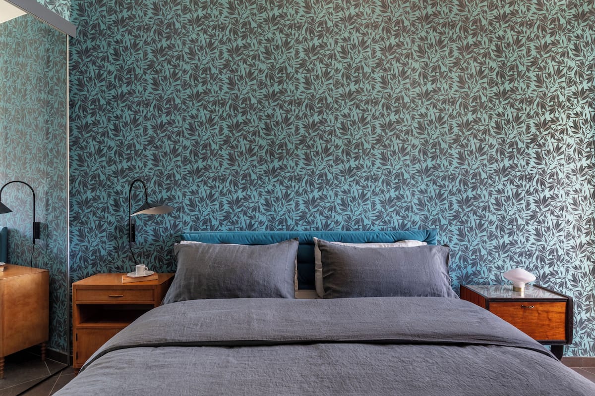 A bedroom with a patterned wallpaper, a bed with grey bedding, two bedside tables, and a nightstand with a lamp.