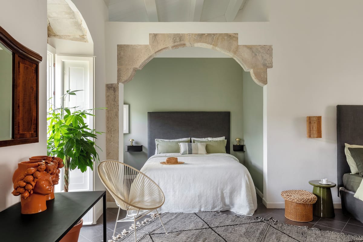 A bedroom with a bed, a chair, a small table, and a potted plant.