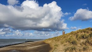 Un paisaje costero con un faro en una duna cubierta de hierba, una playa arenosa y un mar tranquilo bajo un cielo parcialmente nublado.