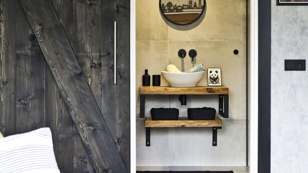 Un baño moderno con una puerta de madera rústica, un espejo redondo, un lavabo sobre encimera y una estantería con toallas y un cuadro de un panda.