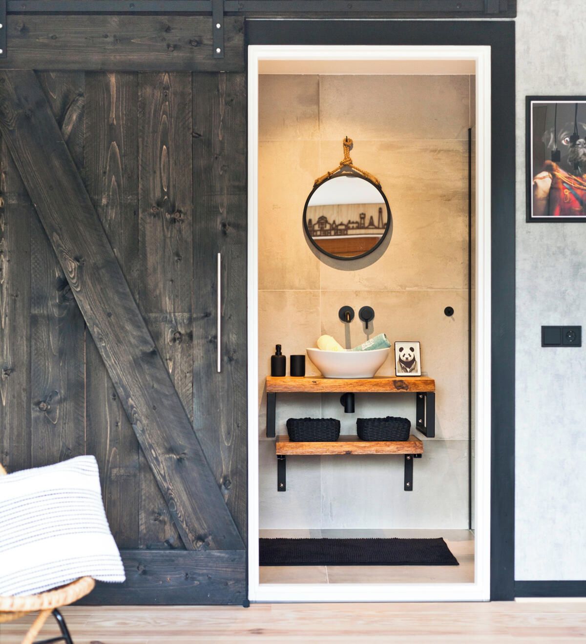 A modern bathroom with a rustic wooden door, a round mirror, a vessel sink, and a shelf with towels and a panda picture.