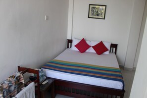 Ein ordentlich gemachtes Bett mit einer gestreiften Tagesdecke, zwei roten Kissen und einem hölzernen Kopfteil. Auf einem kleinen Tisch stehen eine Wasserflasche, eine Tasse und eine Fernbedienung. Ein gerahmtes Bild hängt an der Wand über dem Bett.