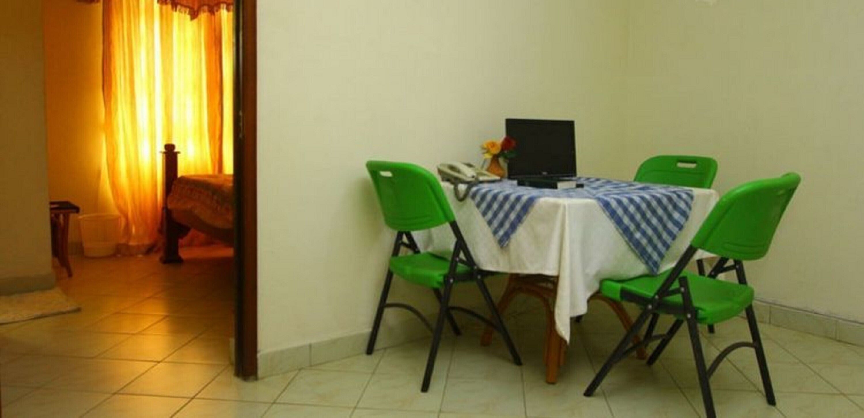 Un petit coin repas avec une table recouverte d’une nappe blanche et des chaises vertes. Un ordinateur portable est posé sur la table, et on peut voir un vase avec des fleurs.