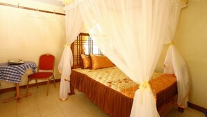 Un dormitorio con cama bajo dosel, una silla, una mesita y una ventana con barrotes.