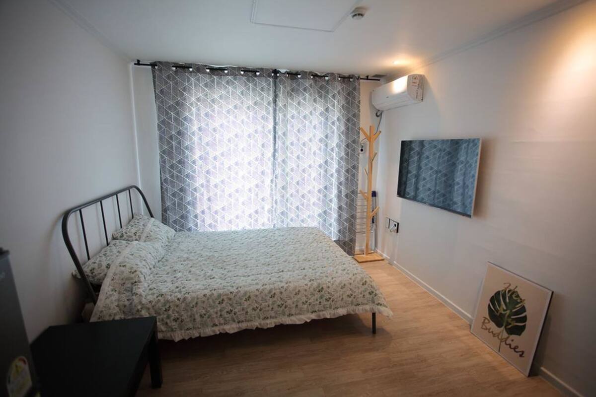 A bedroom with a bed, a bedside table, a wall-mounted air conditioner, a framed picture, and a window with curtains.