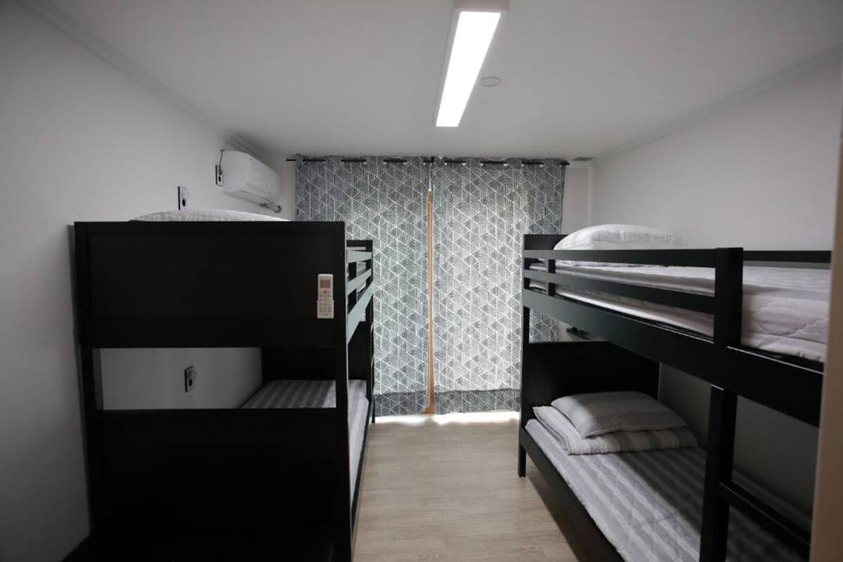 A dormitory room with bunk beds, a window with patterned curtains, and a ceiling light.