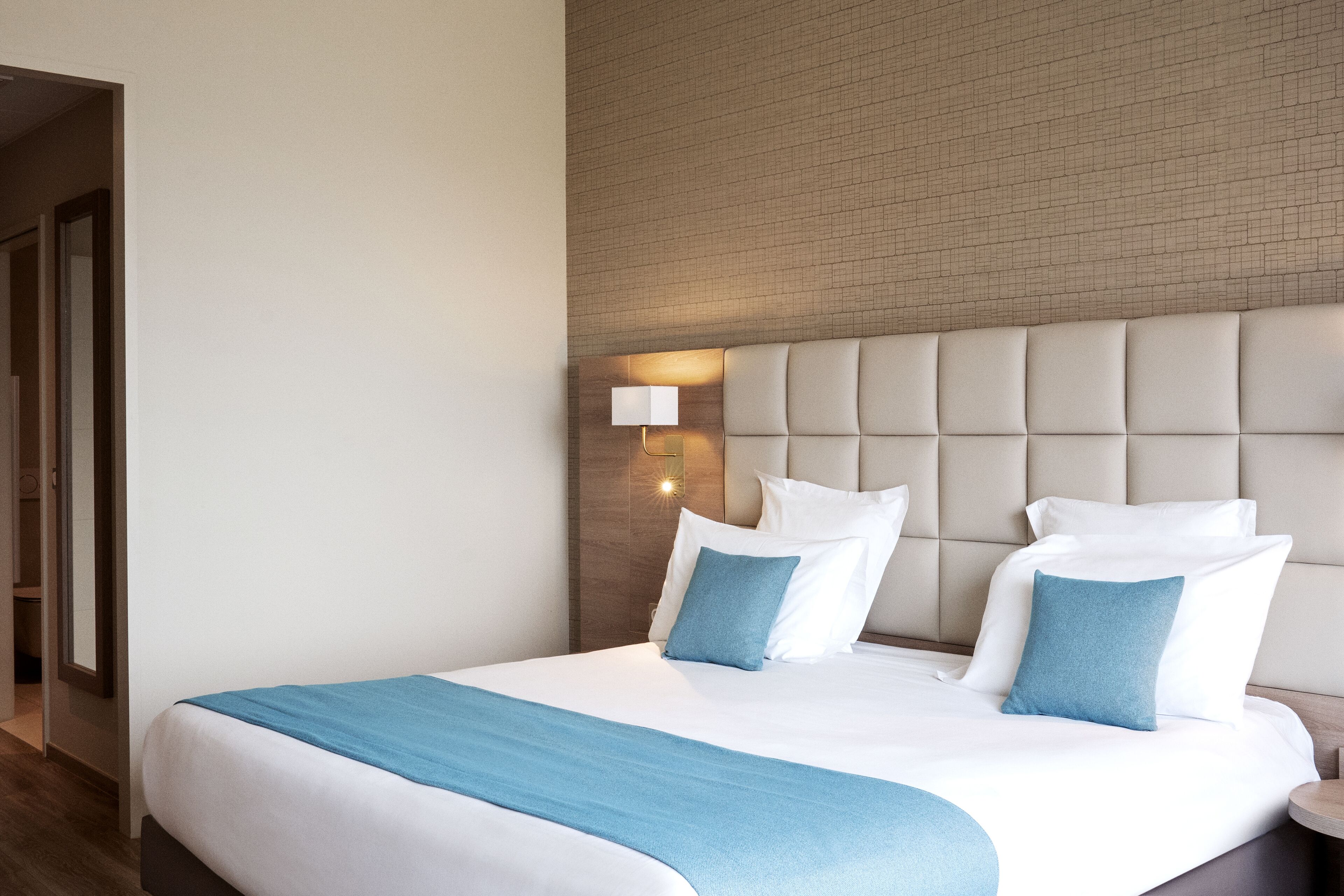 A hotel room with a large bed, a headboard with a light fixture, and a wooden floor.