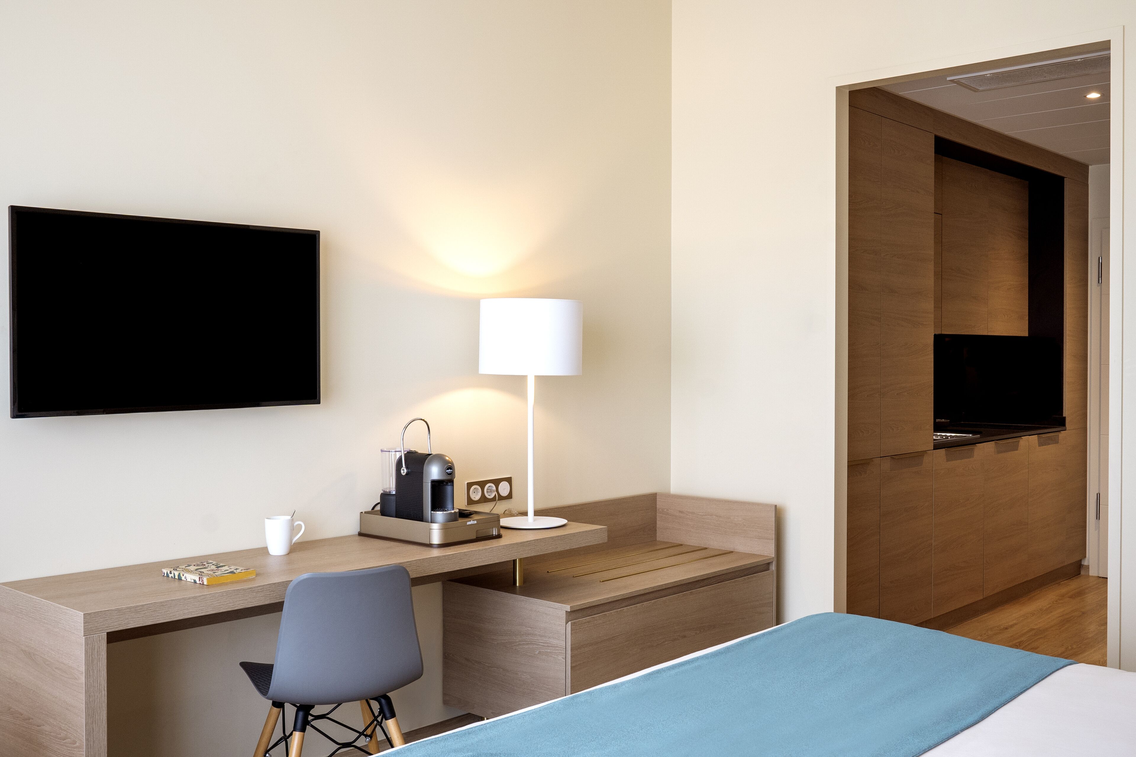 A hotel room with a bed, a desk with a chair, a coffee maker, a lamp, and a TV.