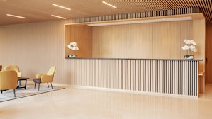 Un área de recepción con un mostrador de madera, dos sillas, una mesita y dos orquídeas en macetas.