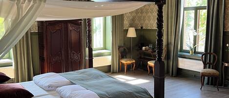 A four-poster canopy bed with a chandelier, a desk with a lamp, and a view of greenery outside.
