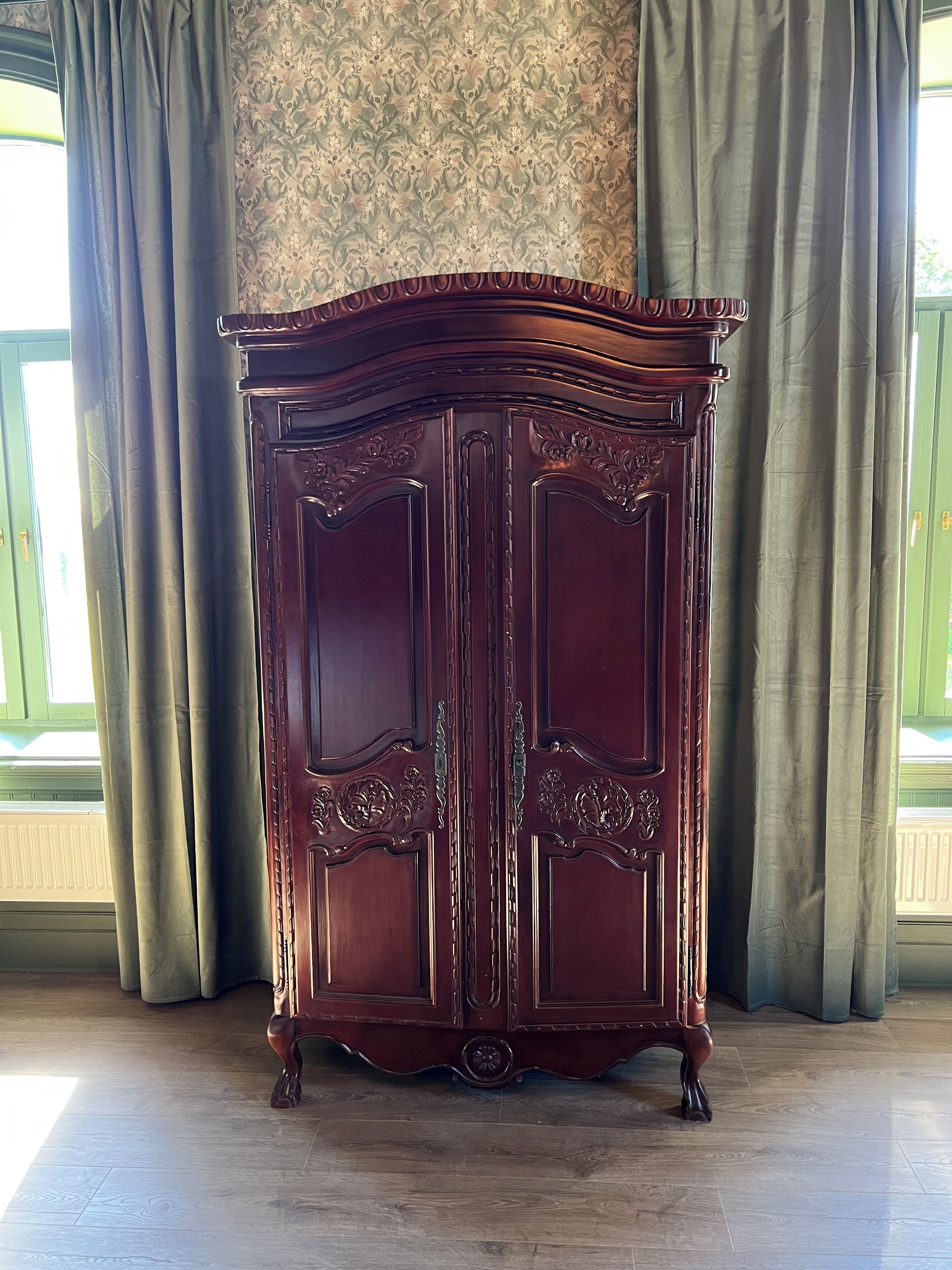 Un meuble en bois sombre, orné de sculptures complexes et surmonté d’un élément décoratif, est placé devant une fenêtre ornée de rideaux verts.