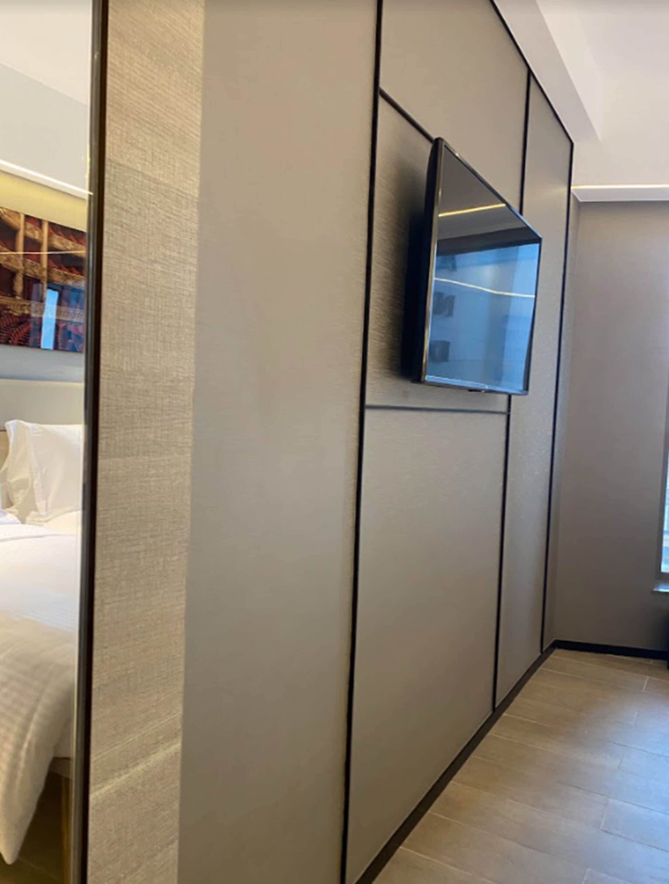 A modern hotel room with a large bed, a flat-screen TV mounted on a wall, and a large window providing natural light.