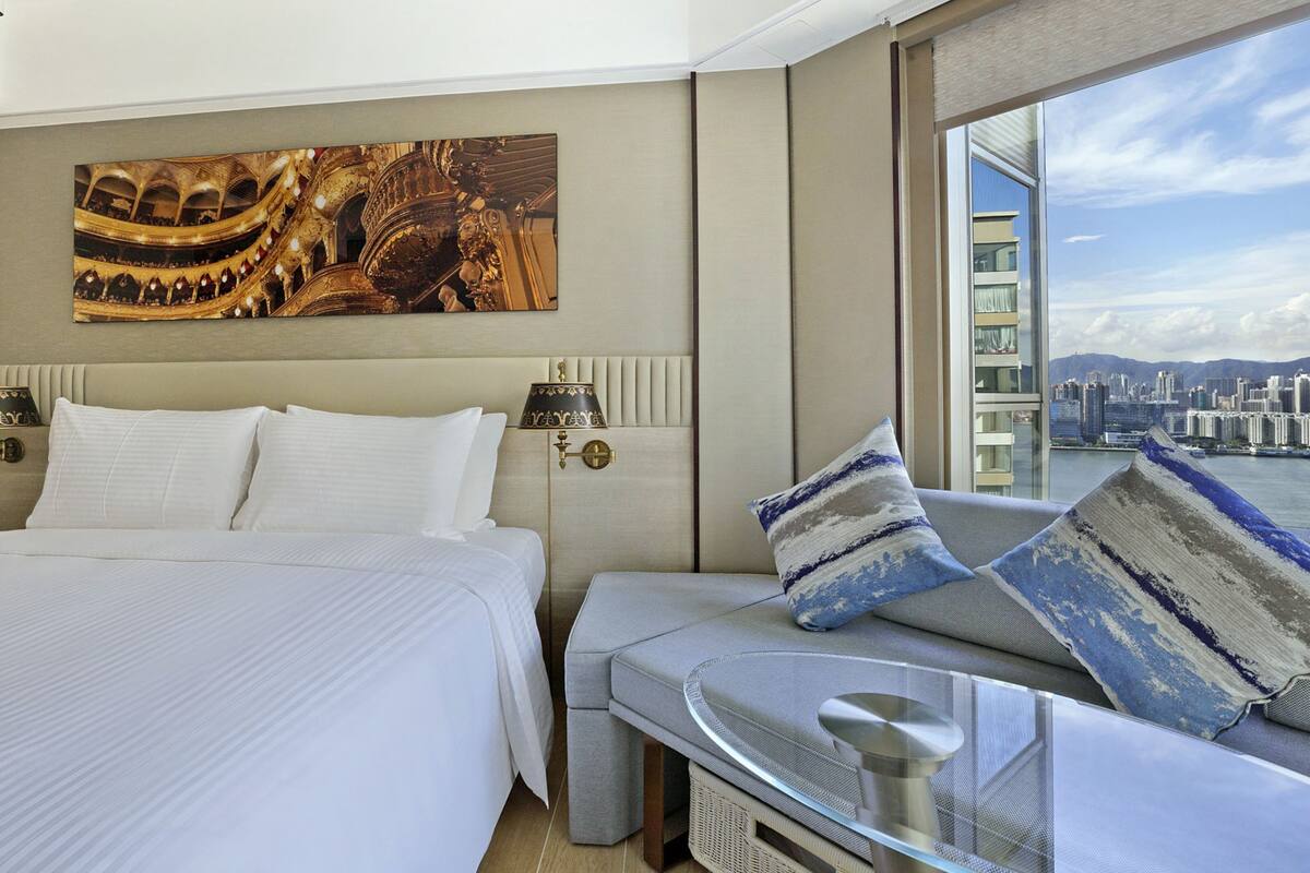 A modern hotel room with a large bed, a sofa with patterned cushions, a glass table, and a city view through a large window.