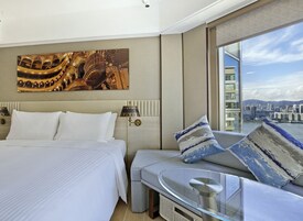 Habitación de hotel moderna con una cama grande, un sofá con cojines estampados, una mesa de vidrio y vistas a la ciudad a través de un ventanal amplio.