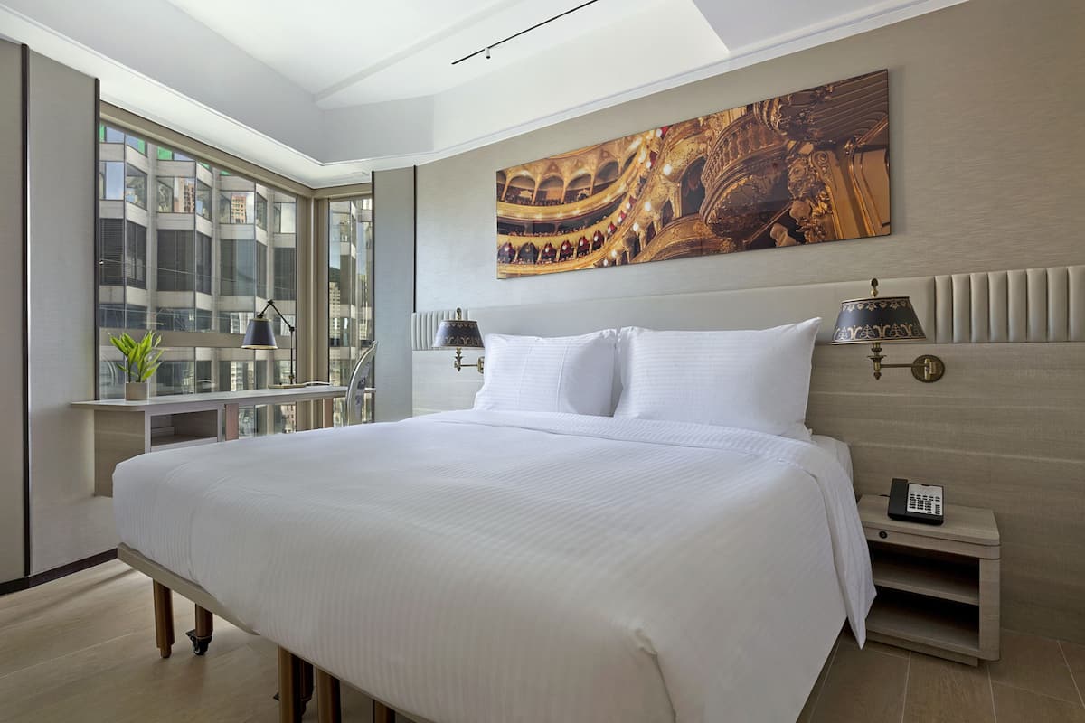 A modern hotel room with a large bed, a view of a cityscape through floor-to-ceiling windows, a painting of an opera house on the wall, and a bedside table with a lamp and phone.