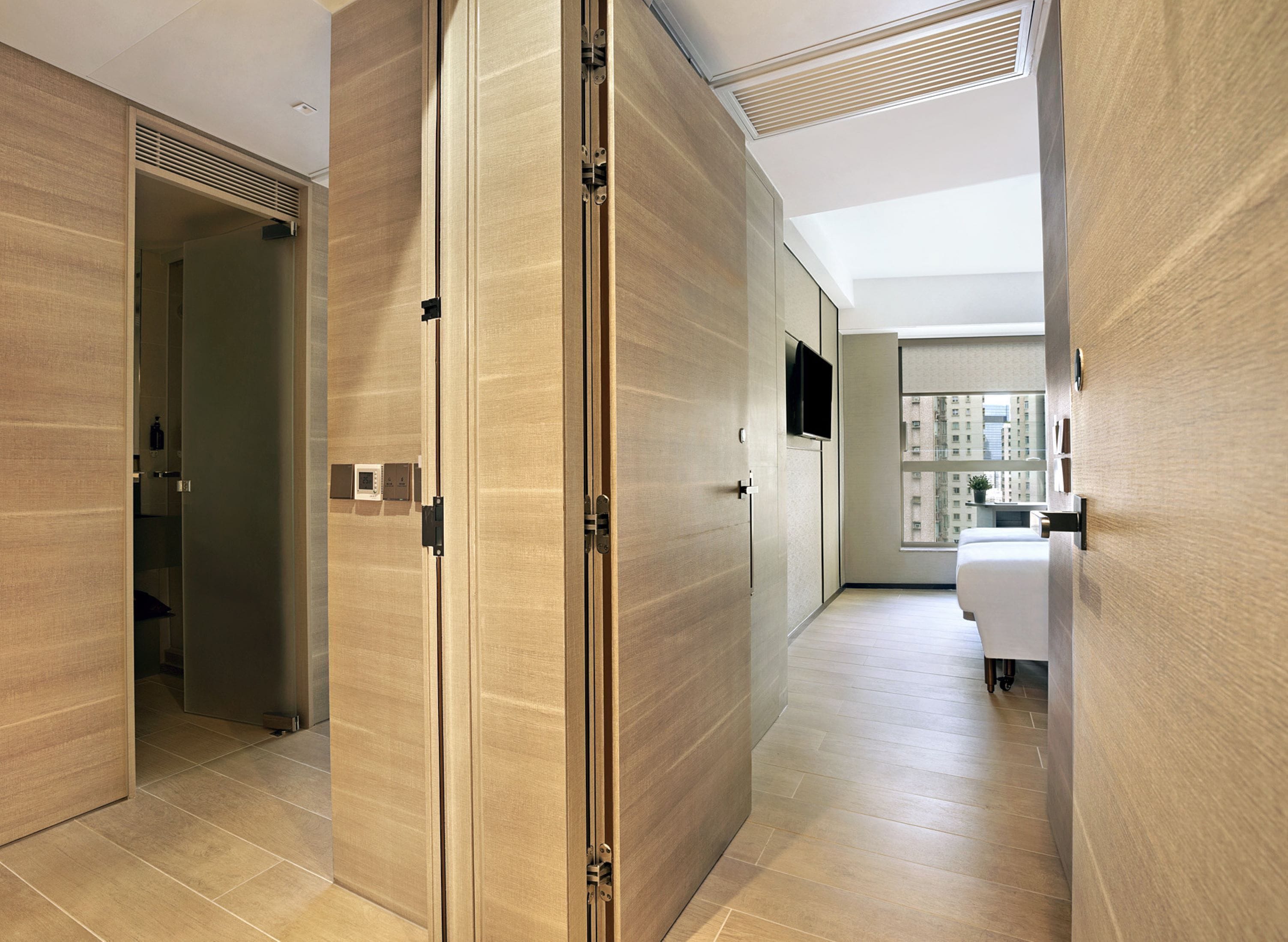 A modern hotel corridor with wooden panel walls, a visible bathroom with a white sink, and a large window offering a city view.